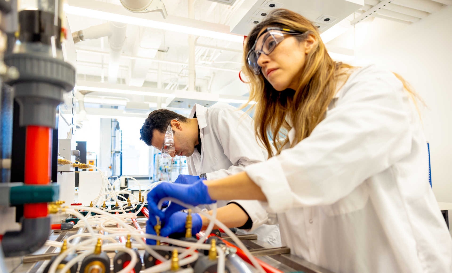 Two students working in a lab, wearing white labs, googles and gloves.