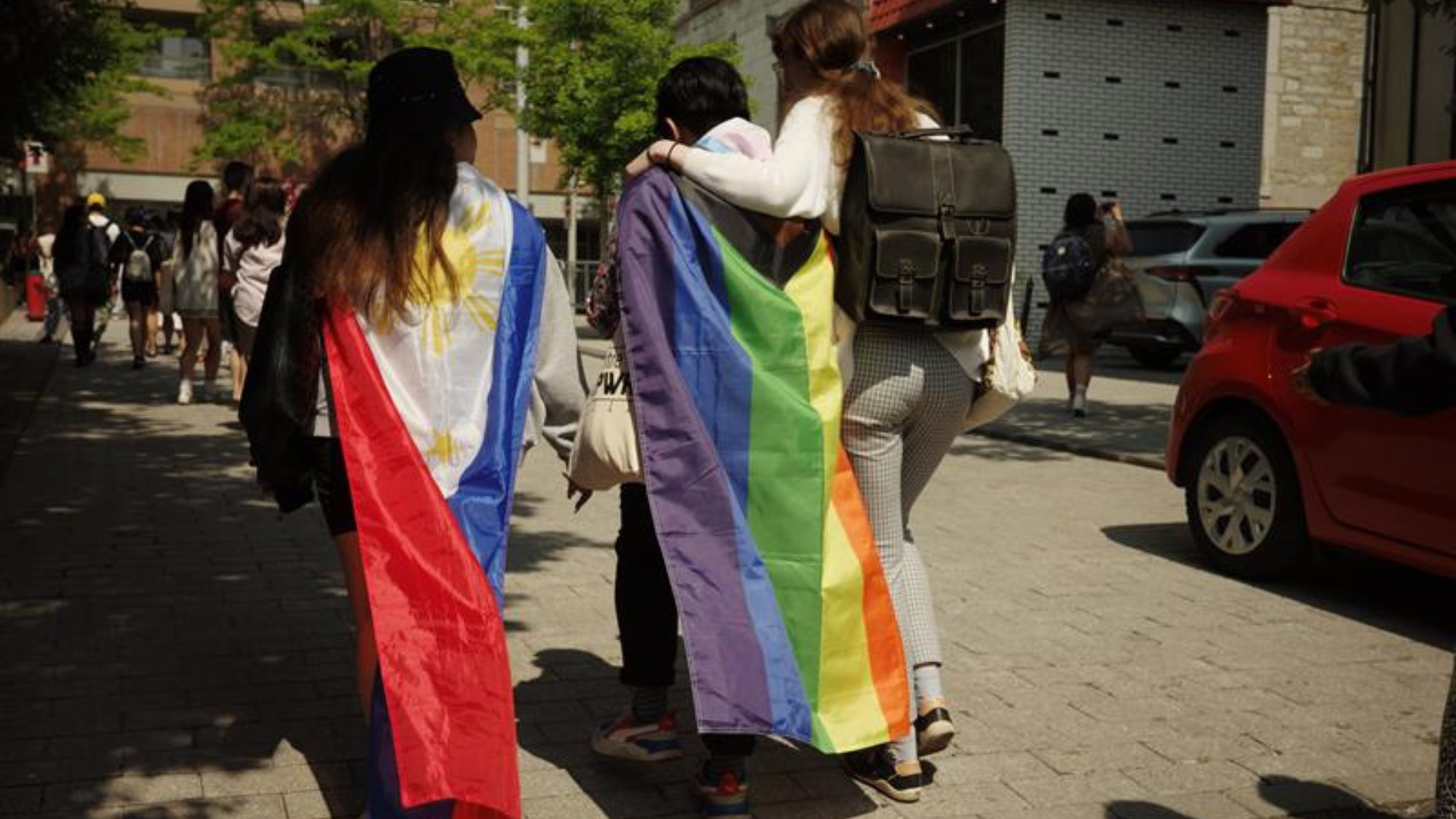 Three people walking away from the camera, one with a Pride flag on their back and another with a Philipines flag
