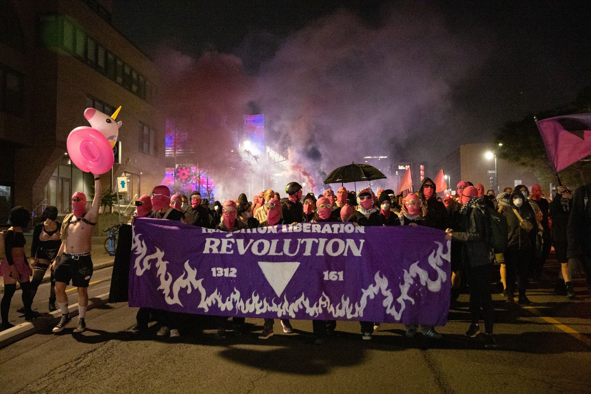 Photograph at night of a group holding a pink banner with white text reading "Queer Revolution Now"