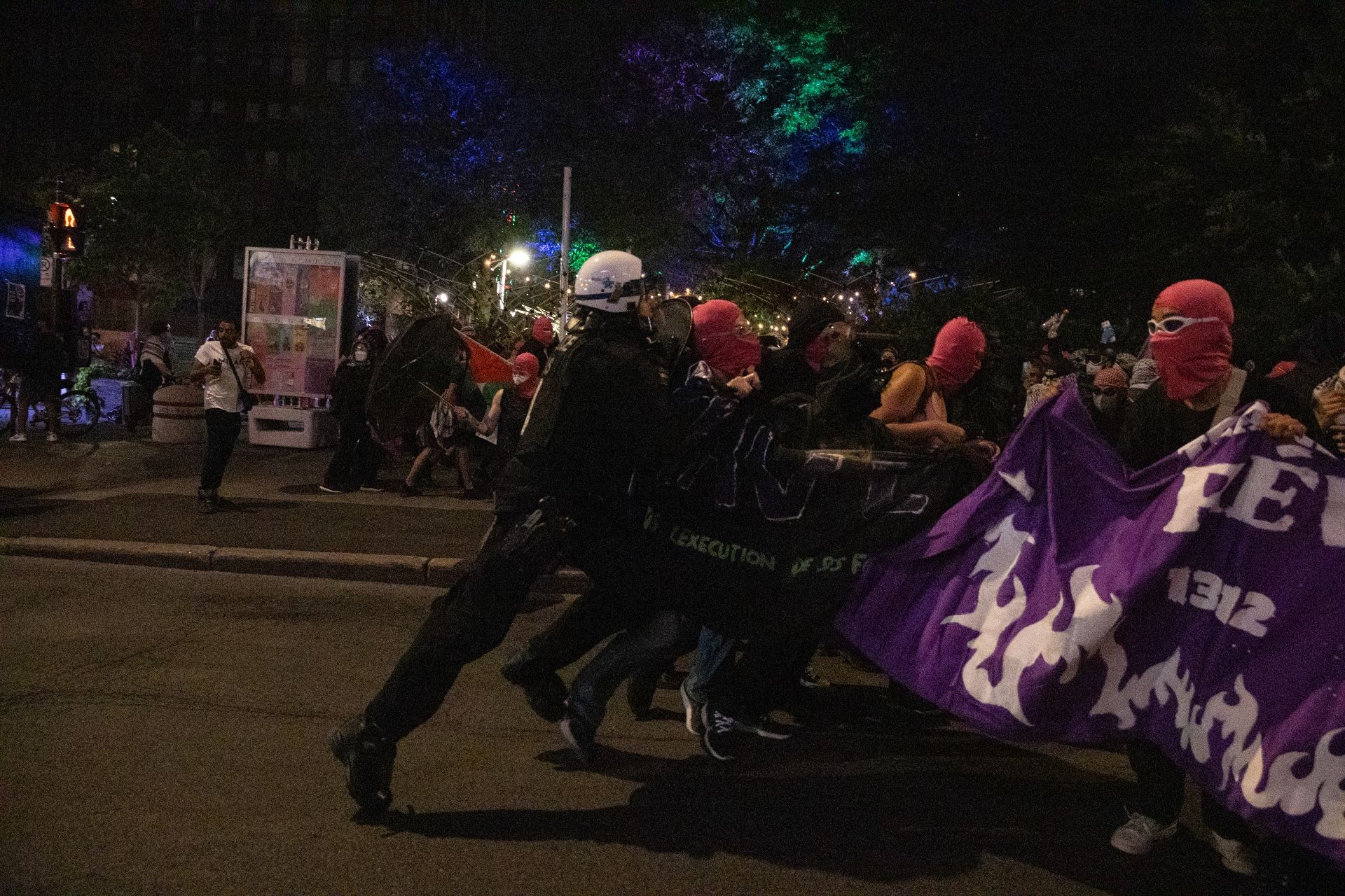 Photograph at night of a group holding a pink banner with white text reading "Queer Revolution Now"