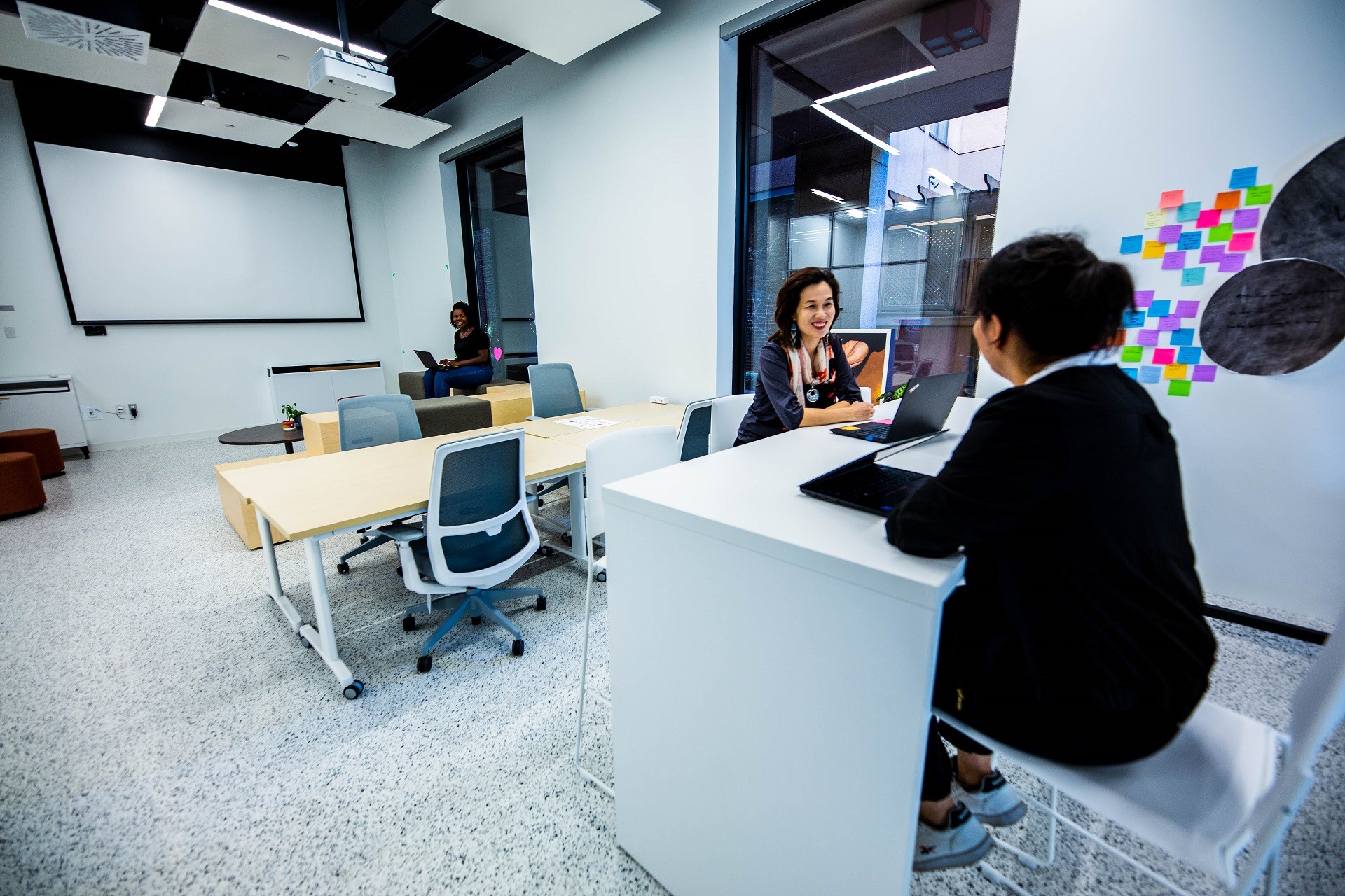 Deux femmes sont assises dans l'espace de co-working, se souriant l'une à l'autre, une autre est vue plus loin dans le fond.