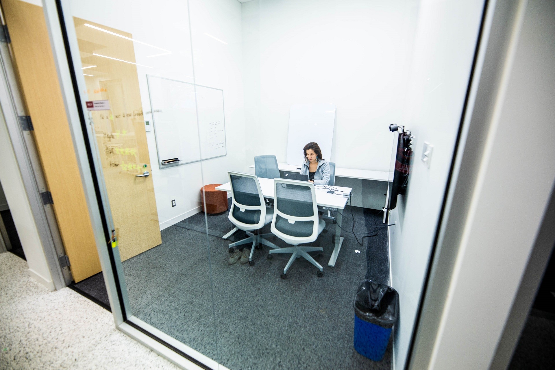 Une personne assise dans la salle de repos et travaillant sur son ordinateur portable. La salle a des murs en verre transparent et comprend une table blanche, un tableau blanc, un écran de télévision et une corbeille. 