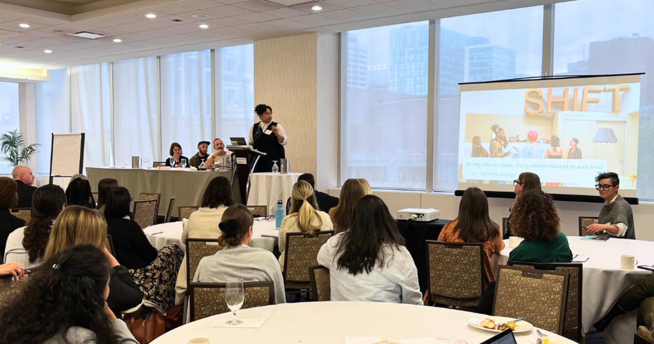 Photograph of an individual standing up on a platform giving a presentation to a room of people, with a slideshow that says "SHIFT" in the background.