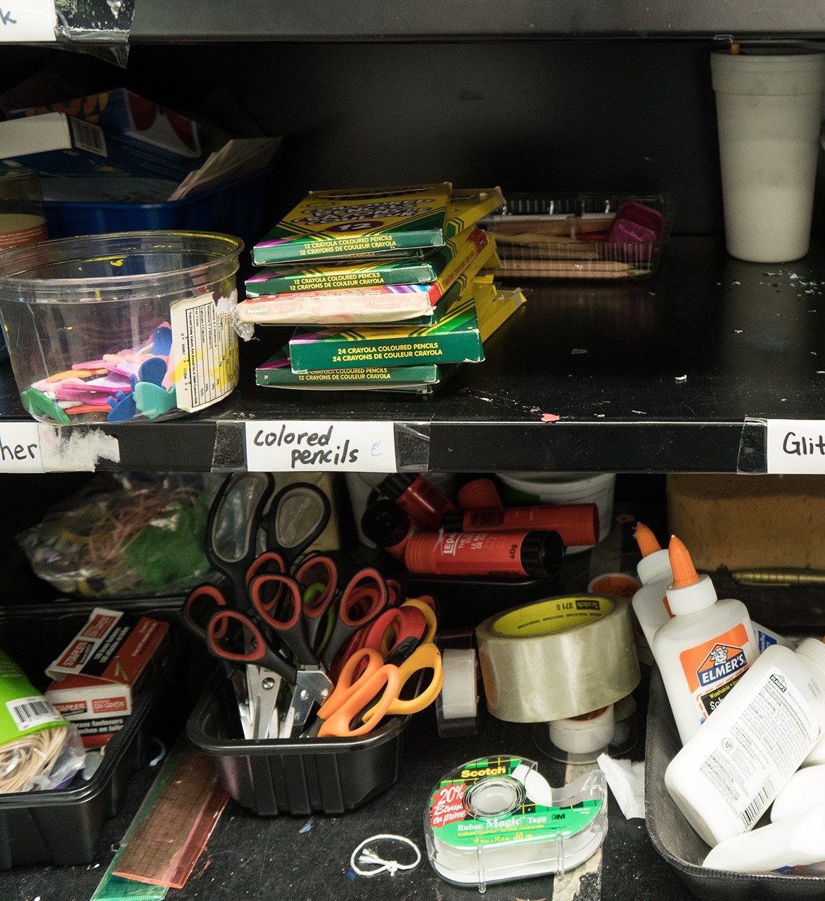 Art supplies on shelves inside a cupboard: container containing stickers, stack of boxes of coloured penciles, scissors, and glue sticks.