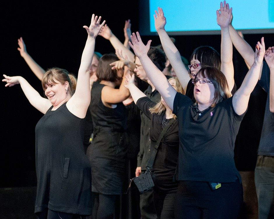 A group of special-needs adults on stage during a performance: they all wear black and have their hands raised over their heads.