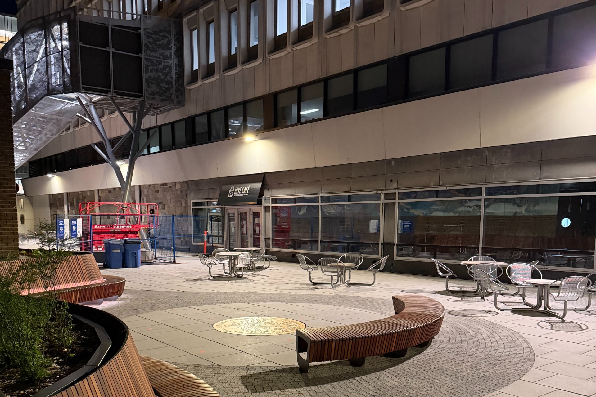 Terrasse rénovée du pavillon Henry-F.-Hall en soirée, avec des bancs en bois incurvés, des tables et des chaises extérieures, des bacs paysagers et l’entrée du café Hive.