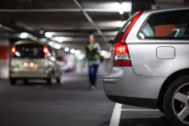 Une voiture de couleur argent est garée dans un stationnement faiblement éclairé. Une personne marche à l’arrière-plan.