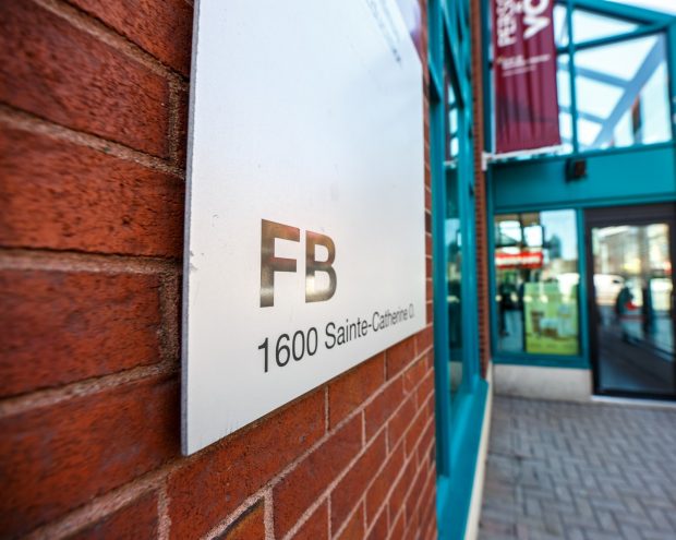 Un panneau sur un mur de briques indique « FB 1600 Sainte-Catherine » près de l'entrée d'un bâtiment à cadre sarcelle avec des portes vitrées et un panneau au-dessus.