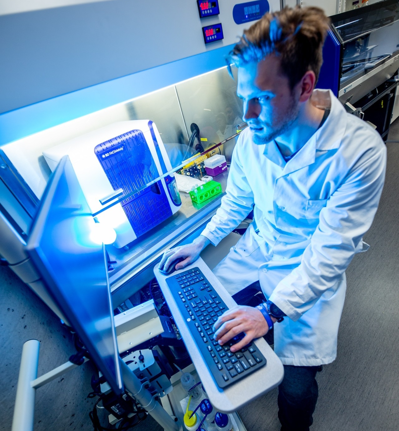 Un scientifique en blouse utilise un clavier d’ordinateur tout en examinant un appareil à éclairage bleu dans un laboratoire.