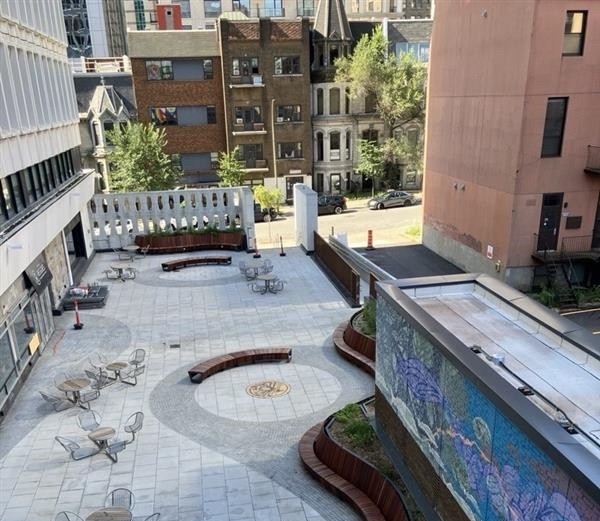 Vue en plongée de la terrasse rénovée du pavillon Henry-F.-Hall, avec des bancs en bois incurvés, des tables et des chaises extérieures, des bacs paysagers, un nouveau pavage en briques et un motif circulaire central.