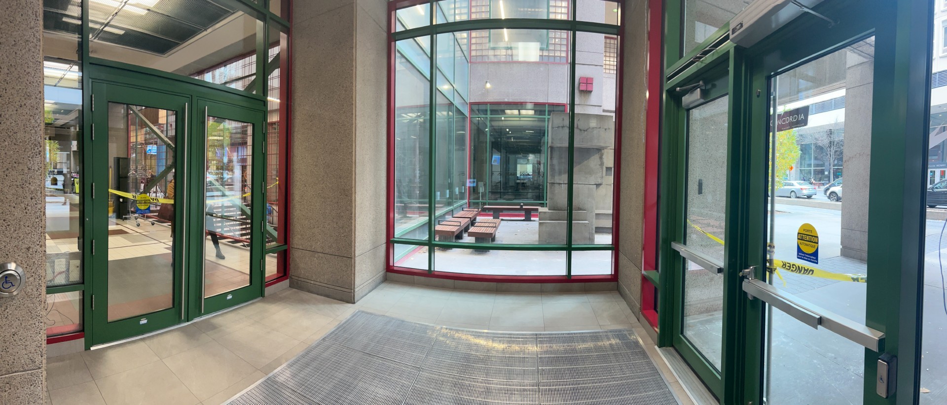 Vue panoramique du vestibule d’entrée rénové du pavillon J.-W.-McConnell (LB), avec des portes vitrées à encadrement vert et de grandes fenêtres donnant sur la cour et ses bancs.