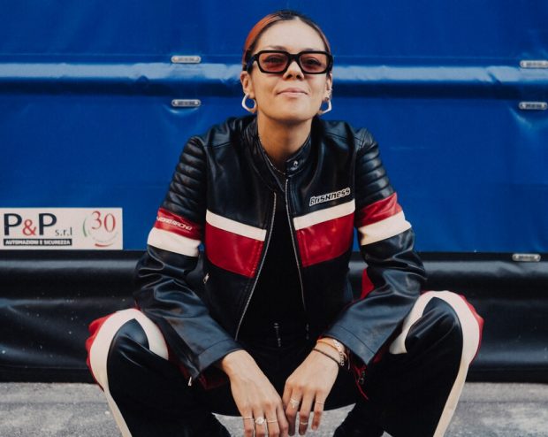 A person in a black leather jacket and sunglasses crouching confidently in front of a blue industrial background.