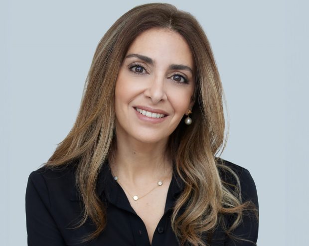 A woman with long wavy hair, wearing a black blouse and smiling against a light blue background.