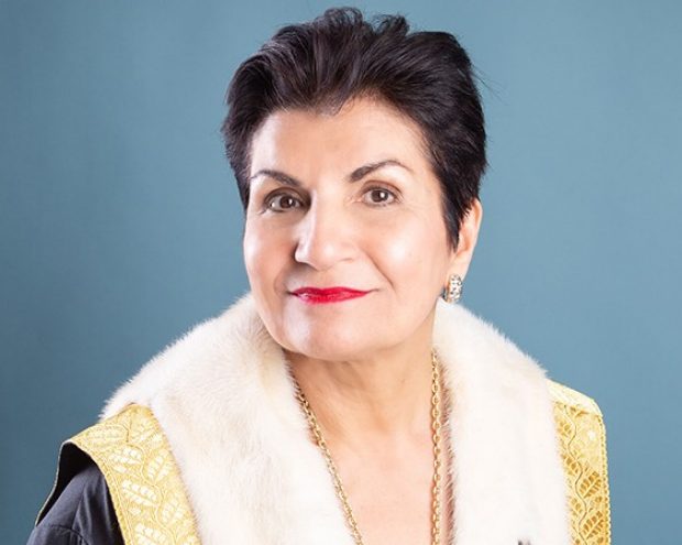 A woman in formal attire with a fur-trimmed robe, smiling warmly in front of a blue backdrop.