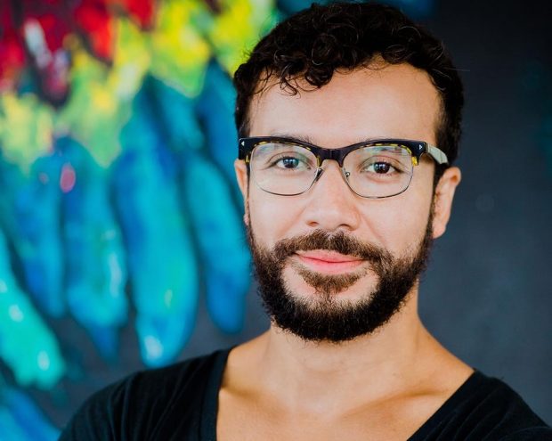 A man with a beard and glasses, wearing a black V-neck shirt, standing against a colorful mural.