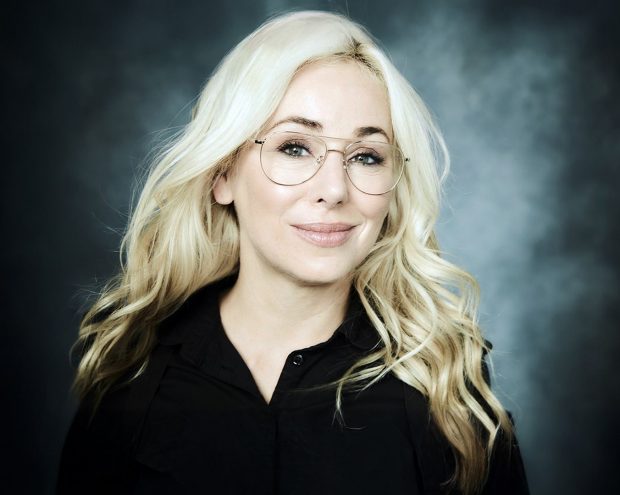 A woman with long blonde hair, wearing glasses and a black blouse, smiling warmly at the camera.