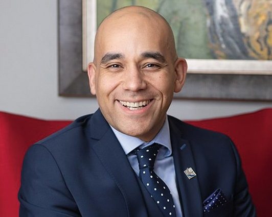A bald man in a navy suit and polka-dot tie, sitting in a red chair and smiling.