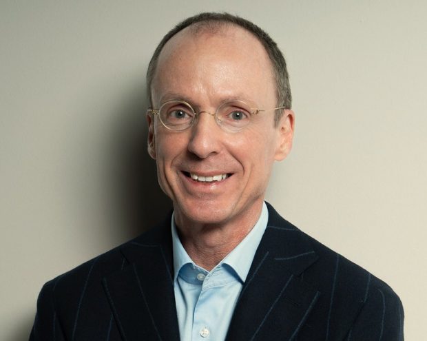 A man in a dark suit and glasses standing with arms crossed in front of a neutral background.