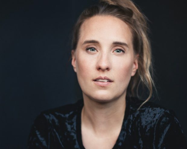 A woman with a bun hairstyle wearing a dark velvet outfit, gazing seriously at the camera.