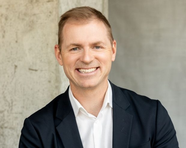A man in a dark suit and white shirt, sitting in front of a concrete wall and smiling broadly.