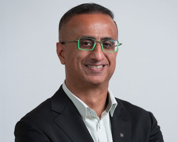 A man with short hair and glasses, standing with arms crossed in a dark blazer against a white background.