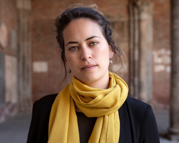 A woman with a yellow scarf and black coat, standing in an old brick corridor with a serious expression.