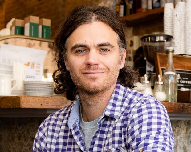 A man with curly brown hair is sitting at a café table smiling at the camera. He is wearing a purple and white checkered shirt