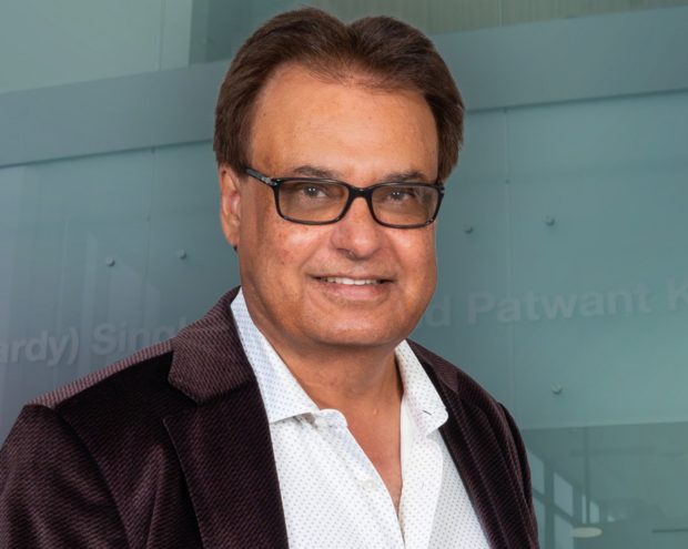  A man in glasses wearing a dark blazer over a white shirt, smiling slightly in front of a glass background.