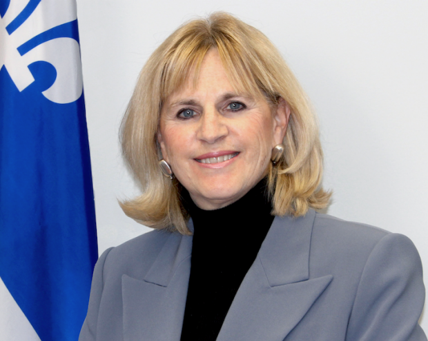 A woman in a grey blazer standing in front of a blue and white flag, smiling warmly.