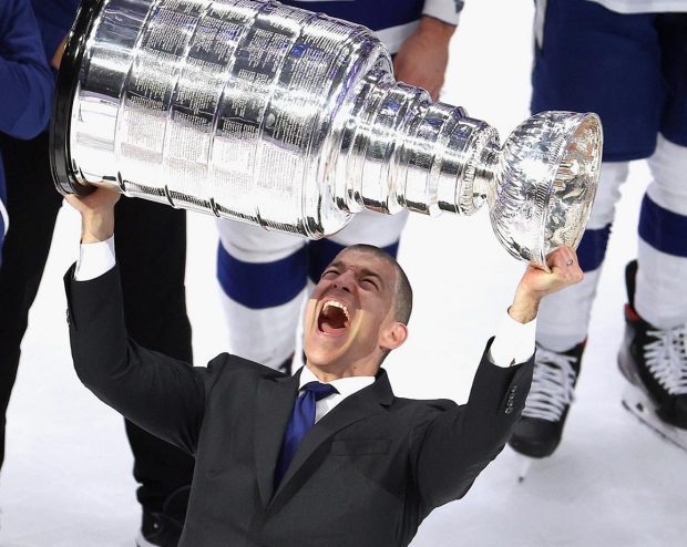 A man in a suit holding a large championship trophy above his head, celebrating on an ice rink.