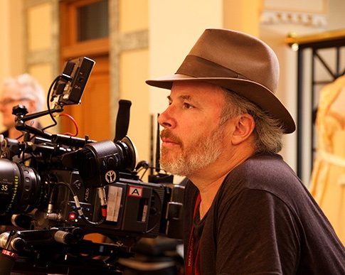 A man with a beard and hat, sitting next to a professional camera setup in a warmly lit indoor space.