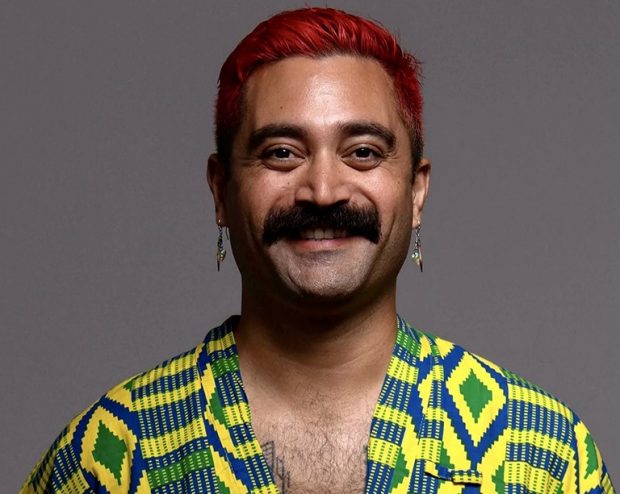 A headshot of a person smiling with short dyed red hair and a moustache wears a yellow and green print shirt