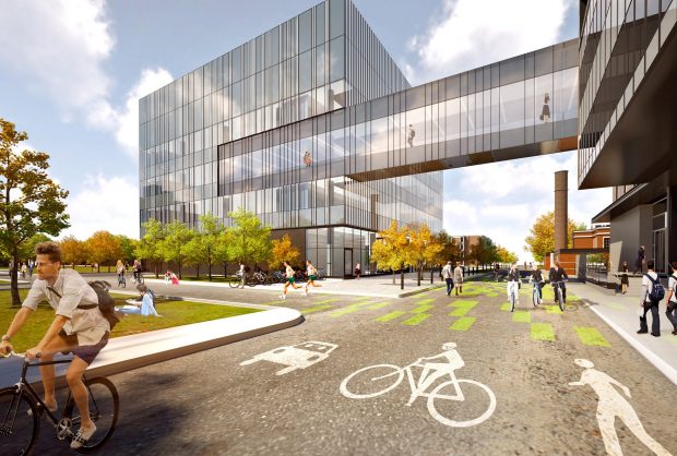 Immeuble de bureaux moderne avec passerelle en verre enjambant une rue dotée de pistes cyclables. Personnes se déplaçant à pied ou à vélo par une journée ensoleillée, sur la route bordée d’arbres.