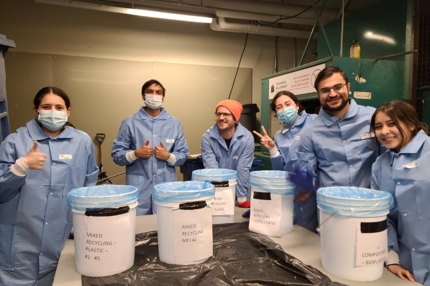 Sept personnes portant des blouses de laboratoire bleues et des masques se tiennent derrière des bacs étiquetés pour le recyclage de divers matériaux, dont le plastique, le métal et le verre. Elles sourient et lèvent le pouce.