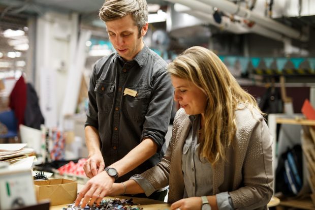 a man and a woman stand at a table sorting small objects against an industrial backdrop