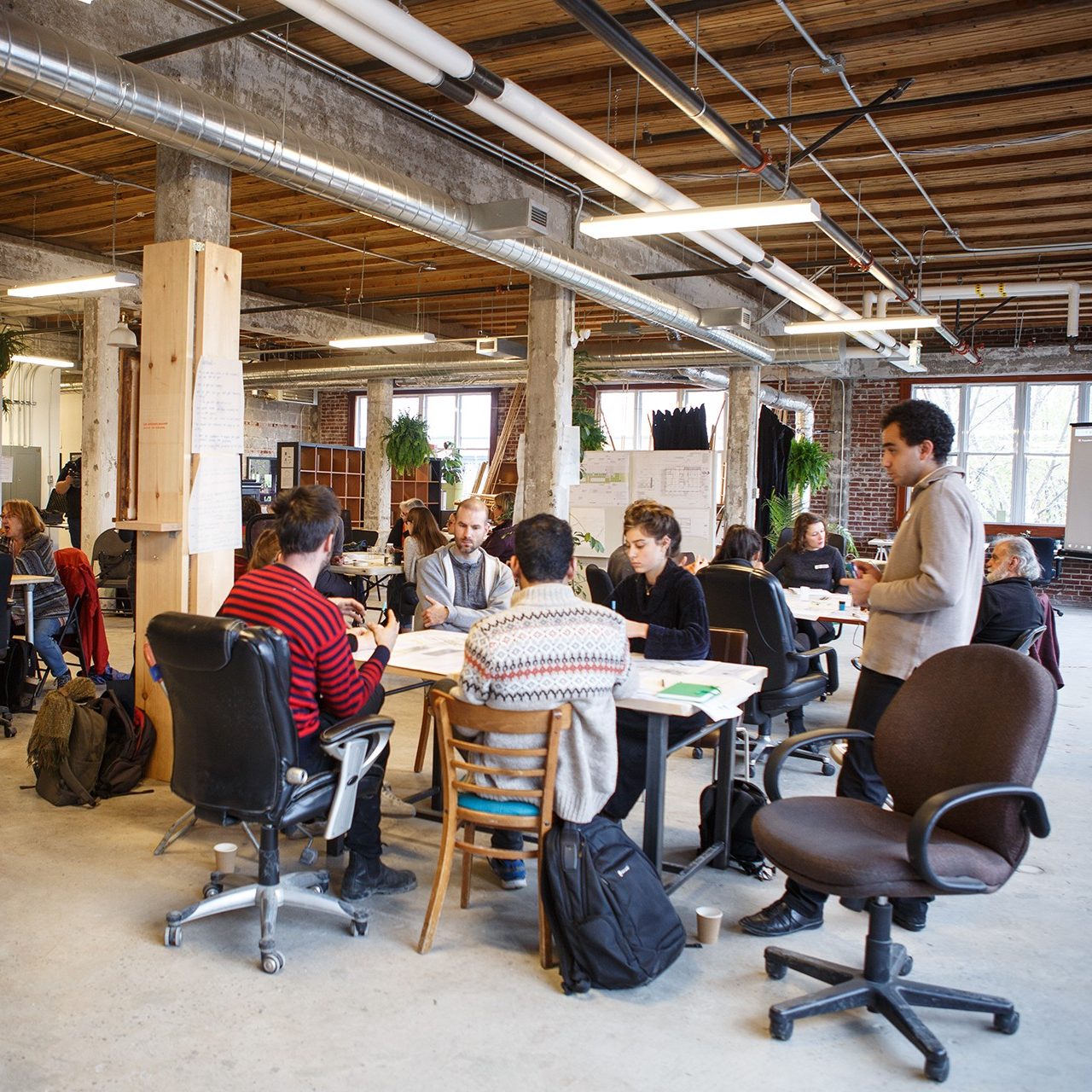 A number of people gather in groups to collaborate around large tables in an open workshop space with exposed beeams, ceilings, and brick walls. Plants and whiteboards are dotted throughout the space.