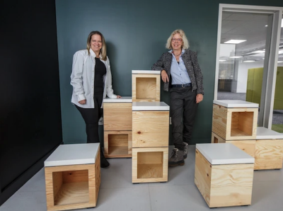 Deux femmes souriantes se tiennent dans une pièce parmi des meubles de bureau modernes.
