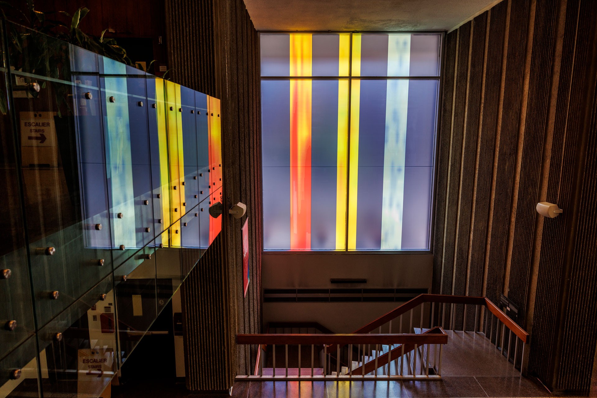 Vue intérieure d'une cage d'escalier dans un immeuble. De grands panneaux verticaux translucides aux couleurs vives (jaune, rouge, bleu et orange) remplissent une fenêtre en haut de l'escalier, projetant une lumière colorée et des reflets dans l'espace.