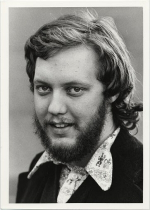  Black and white portrait of a man with a beard and medium-length hair, wearing a patterned shirt and a jacket.
