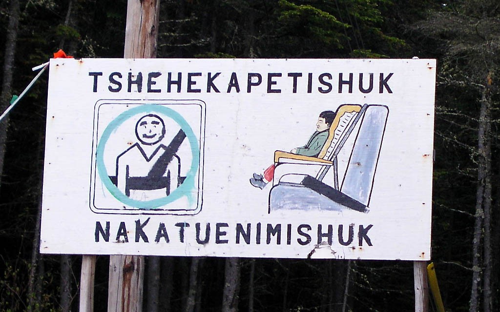 Un panneau « Port obligatoire de la ceinture de sécurité » peint à la main, avec l'inscription d'une traduction en innu-aimun « Tshehekapetishuk nakatuenimishuk » | A hand-painted “Mandatory seat belt use” sign, with the Innu-aimun translation “Tshehekapetishuk nakatuenimishuk”