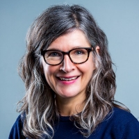 Une femme avec des lunettes et des cheveux grisonnants porte un pull bleu marine.