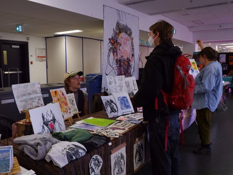 Une personne portant un masque et un sac à dos regarde des œuvres d'art exposées sur une table dans un marché couvert ou une foire d'art. Une autre personne lui parle depuis l'arrière de la table.