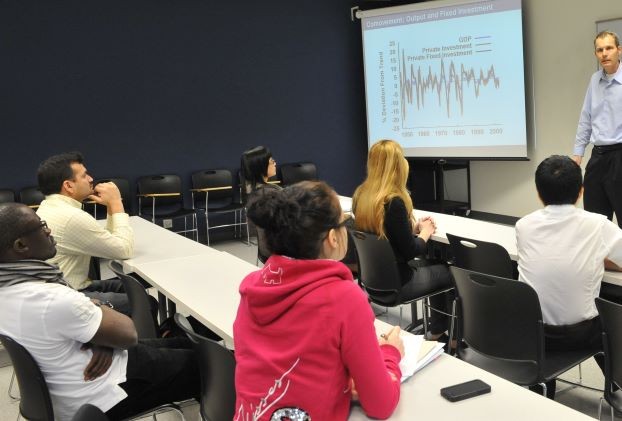 Five students seated at two rows of desks in a classrom, with a male instructor at the front, explaining a chart projected onto a screen.