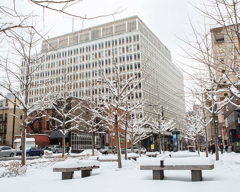 A winter scene depicting a large white campus building with snowy trees in the foreground