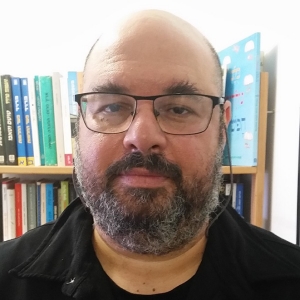 Man with glasses and black shirt with bookcase in background