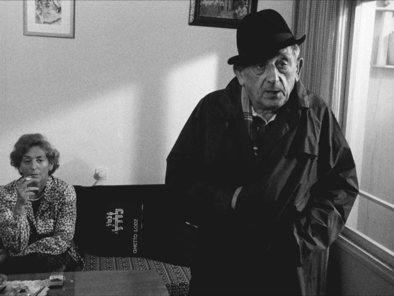 Black-and-white indoor photo of standing man wearing hat and dark coat with sitting woman smoking cigarette