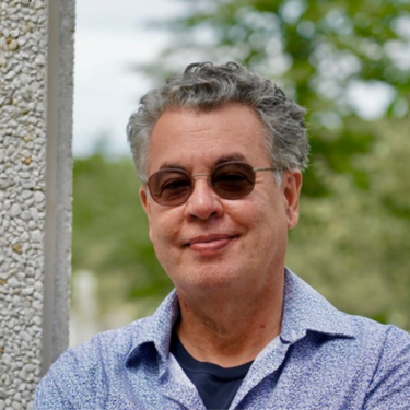 Man wearing brown sunglasses and collared light-blue shirt smiling with mouth closed outdoors