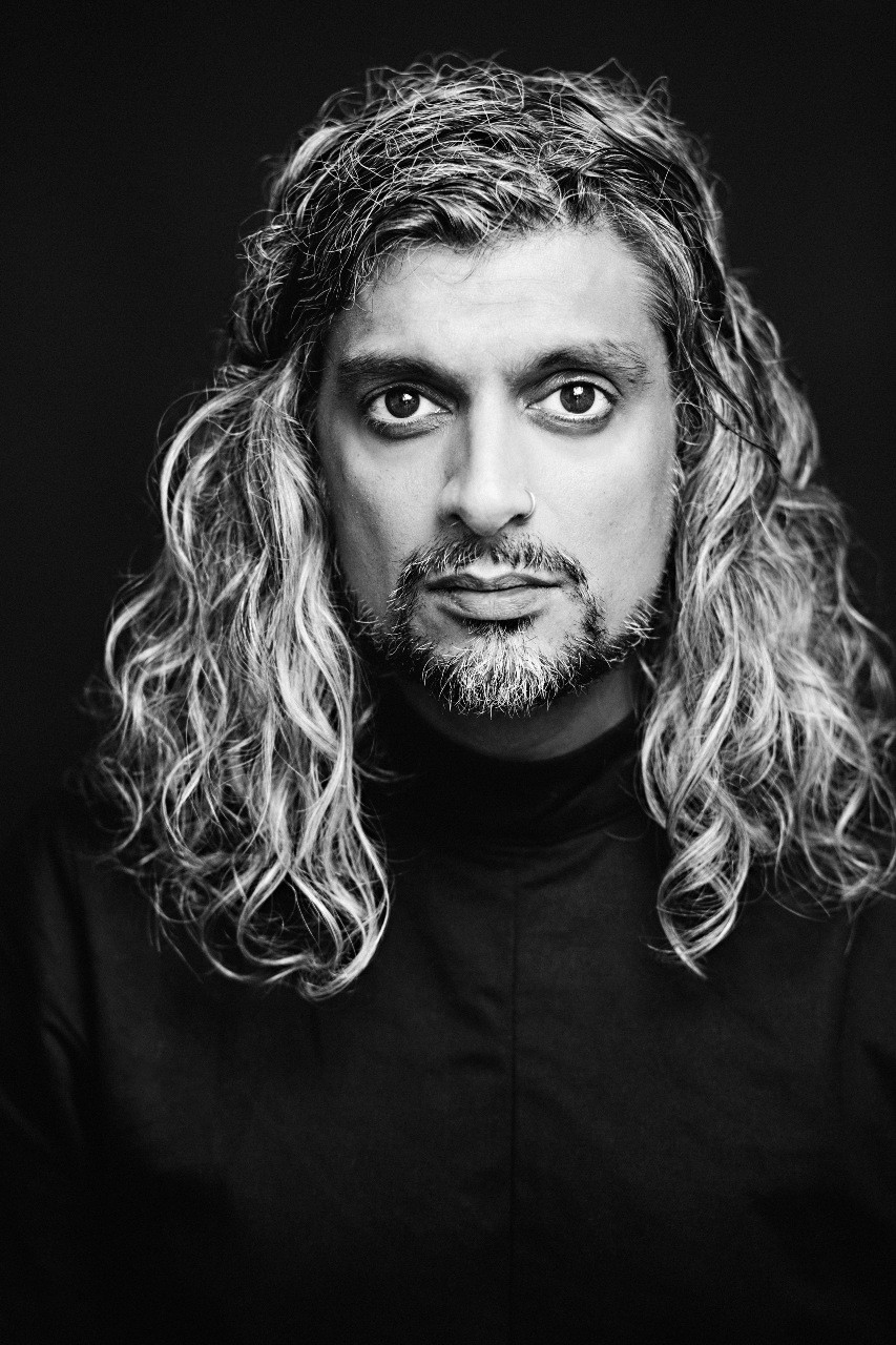 Black and white portrait of Kazim Ali. He has long curly hair and a goatee.