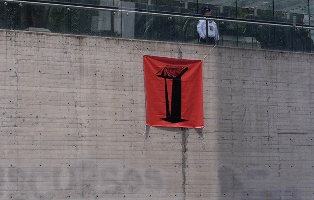 Red banner with a black podium logo hanging on a large concrete wall with a security agent in the background. 