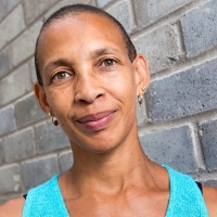 A photo of a blue-eyed, bronze-skinned woman wearing a turquoise tank top against a background of grey bricks
